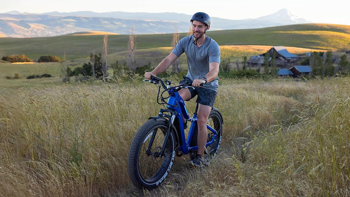 OKAI Ranger e-bike being ridden by middle aged man across a field with tall grass within post for Lectric XPedition dual-battery cargo e-bike