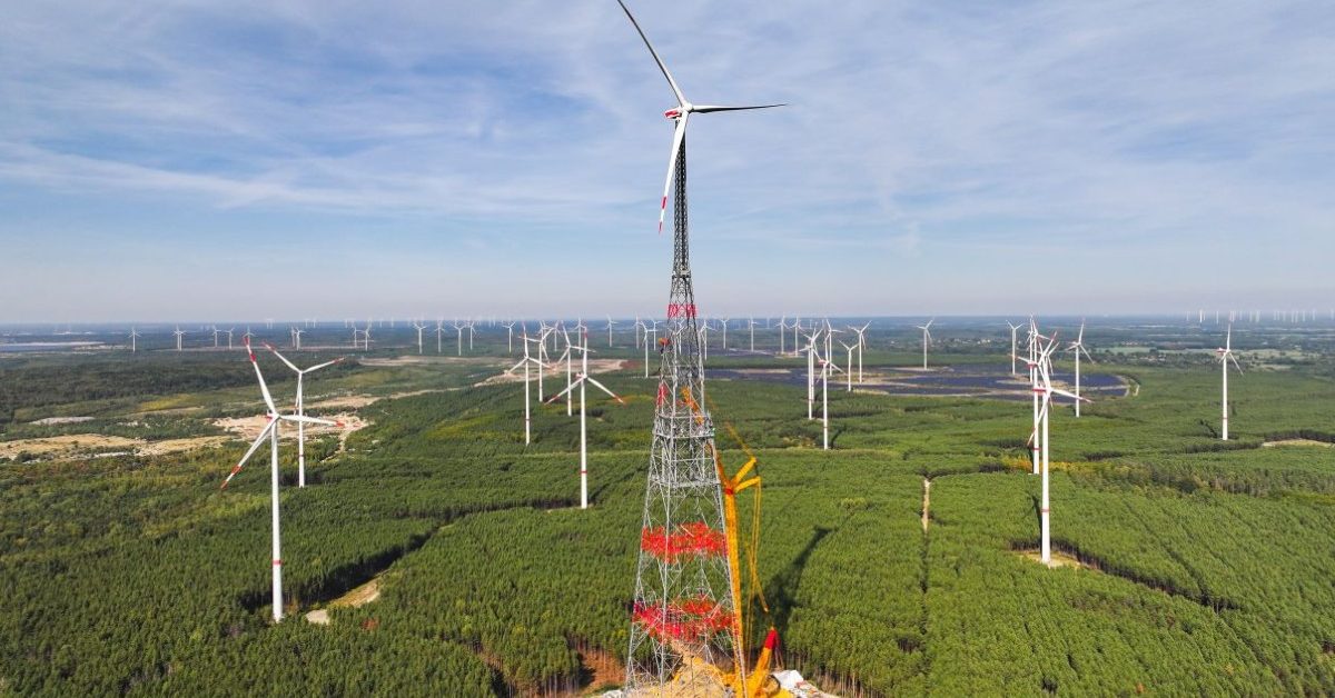 A towering wind turbine with a lattice structure and telescoping tower stands against a sky in Schipkau, Germany.