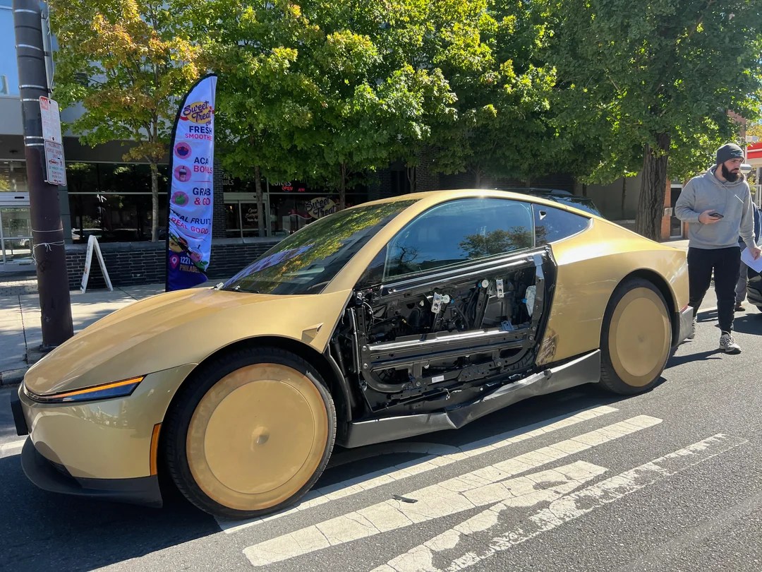 Gold Tesla Cybercab stopped on South Broad Street in Philadelphia with the outer panel of its driver-side door missing